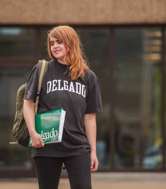 A young female Delgado student wearing a Delgado t-shirt poses in front of an academic building holding a Delgado branded folder and wearing a backpack.