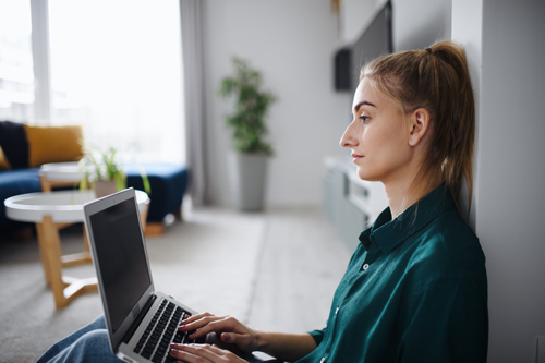 white female student on a computer leaning against a room wall
