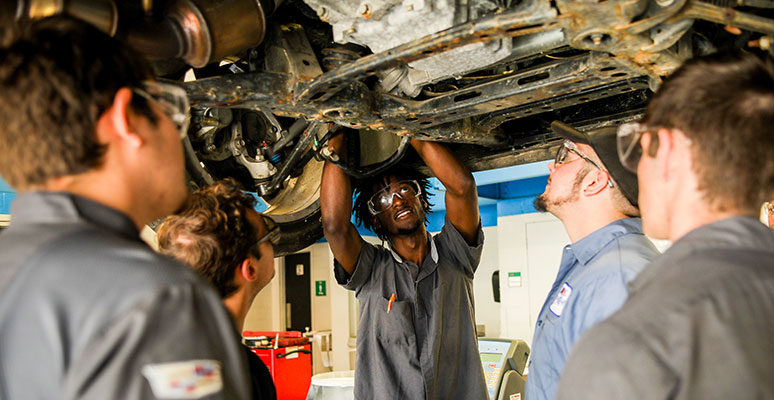 student working on automobile while others are watching