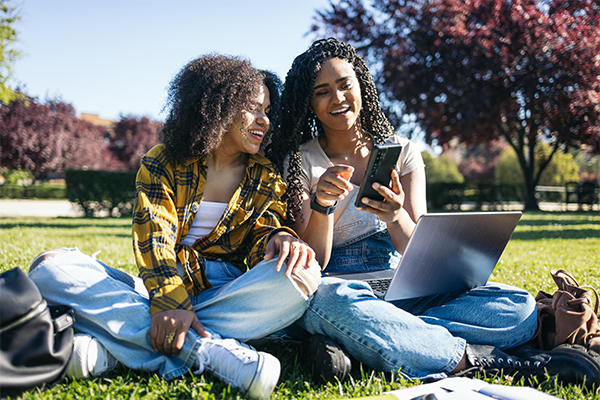 Two students sitting on campus look at their phone and laptop