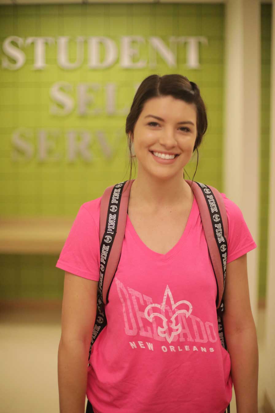 A Delgado student wearing a pink shirt smiles in front of a wall that says "Student Self Service"